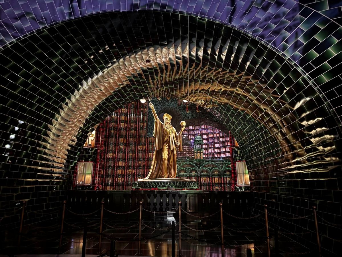 A golden statue of a robed figure stands under a dark, arched tunnel lined with glossy, dark green tiles that reflect the statue and surrounding light.